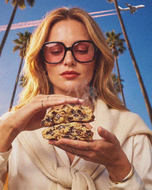 Une jeune femme tient un cookie maison Woody's Factory coupé en deux, encore chaud, sous les palmiers