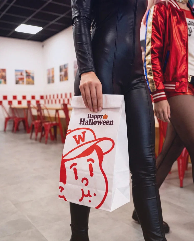 Sac en papier Woody's à la mascotte, avec un sticker Happy Halloween, tenu par une personne en combinaison noire dans une salle aux chaises rouges et au damier rouge et blanc