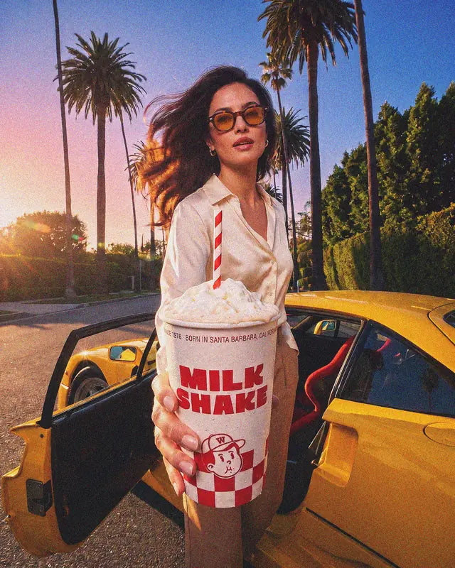 Une jeune femme tend son milkshake Woody's Factory devant une voiture jaune, sous les palmiers au coucher du soleil