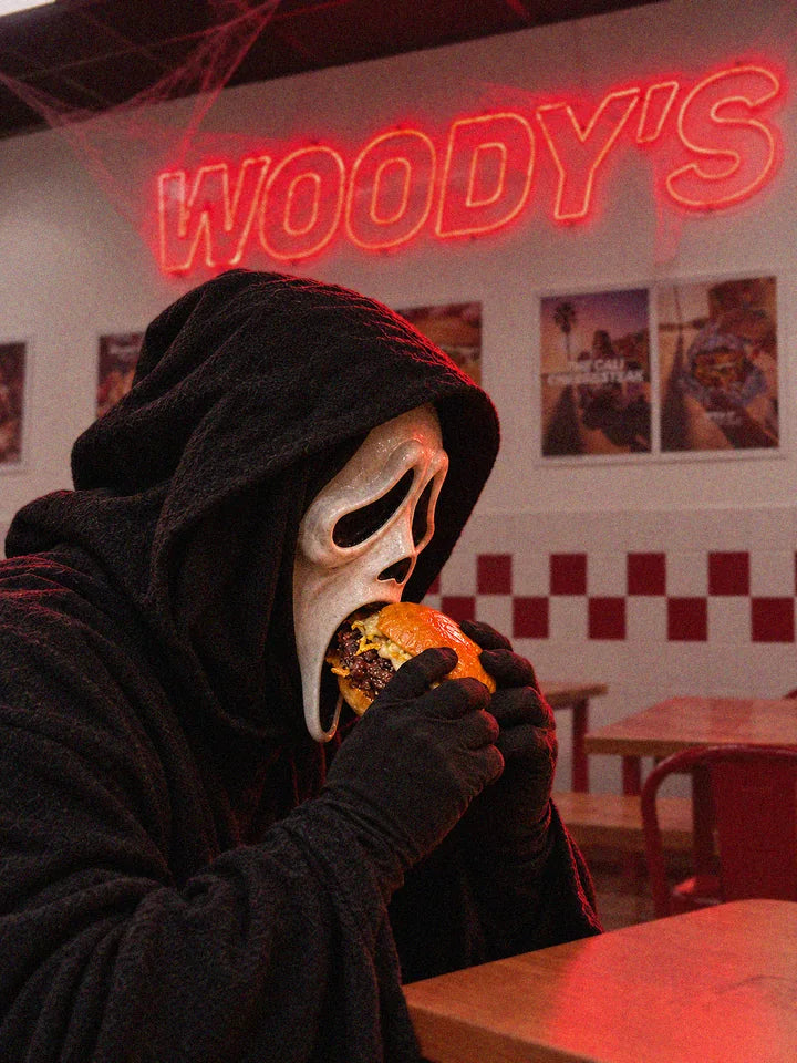 Soirée Halloween chez Woody's : un convive déguisé, capuche noire et masque blanc, croque un smash burger sous le néon Woody's